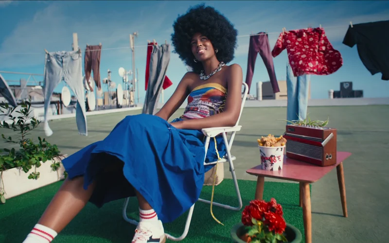 A woman in a blue skirt sits on a rooftop beside a KFC bucket in Feed Your Original
