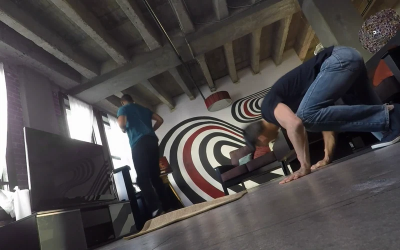 A man balances on his hands beside a black-and-white swirl mural in Like