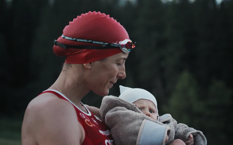 Nicola Spirig in a red swim cap holds her baby beside an alpine lake
