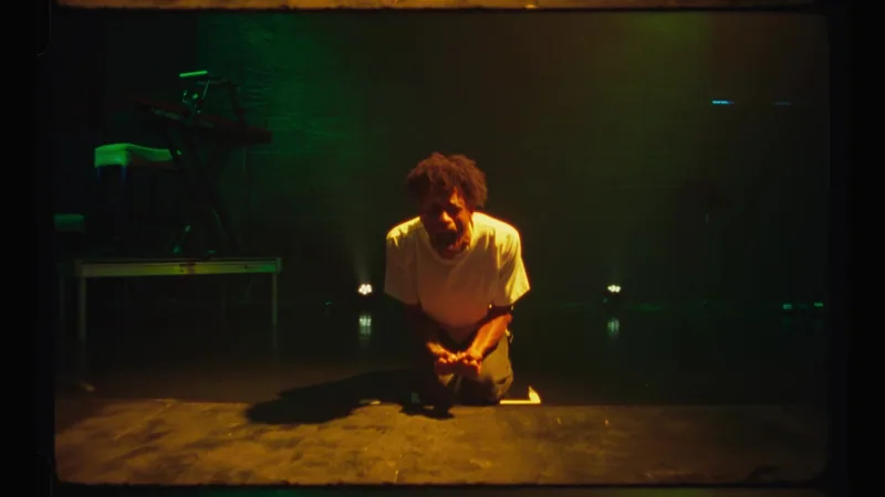 A performer in a yellow shirt leans over a desk on a dark theatre stage.