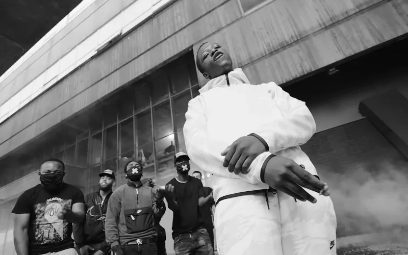 Pa Salieu in a white tracksuit performs with friends beneath a concrete walkway in black and white.