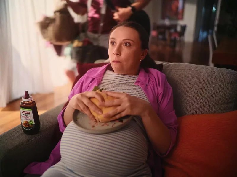 A pregnant woman holds a burger while seated on a sofa.