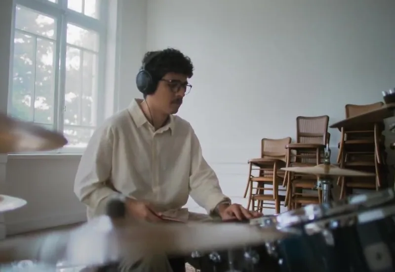 A drummer sits at a drum kit beside a bright window.