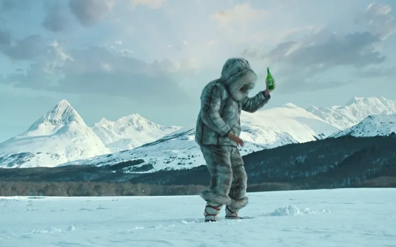 A dancer in a fur-trimmed snow suit holds a green bottle against a snowy mountain backdrop.