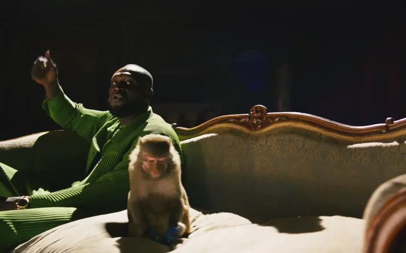 Stormzy in a green suit seated beside a monkey on an ornate sofa.