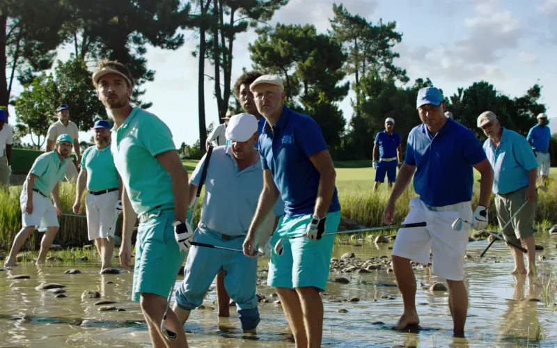 A crowd of golfers in blue and green clothing wades through a pond.