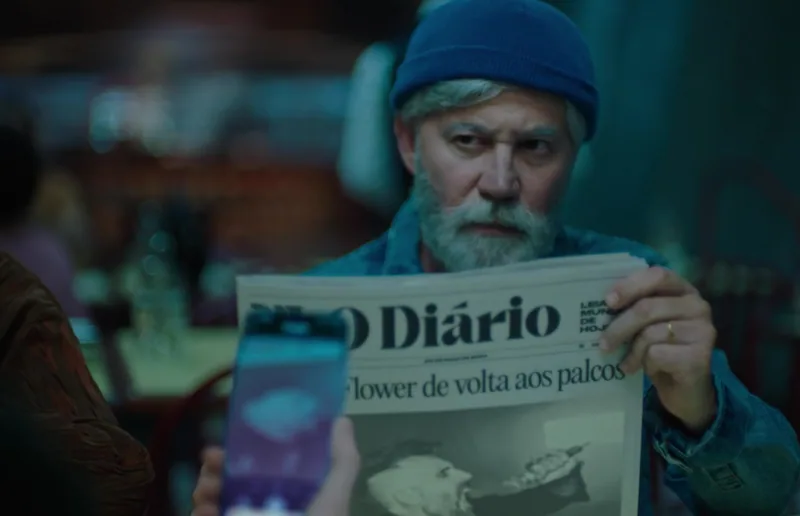 A bearded man reads a newspaper with a phone in the foreground.