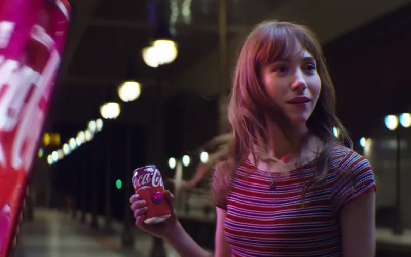 A woman holds a Cherry Coke on a station platform beside a red vending machine.