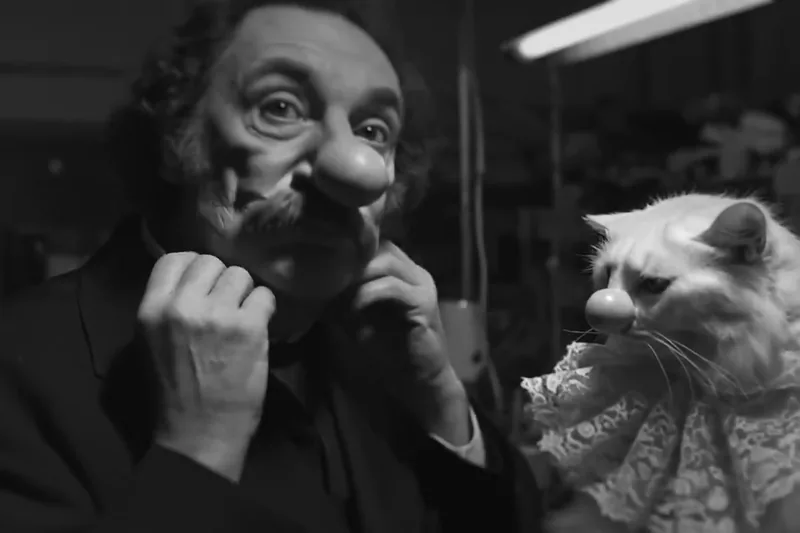 An expressive moustached man holds a cat in a monochrome scene.