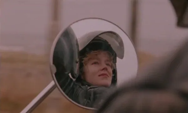 A woman wearing a motorcycle helmet is reflected in a round mirror in Blue Jean.