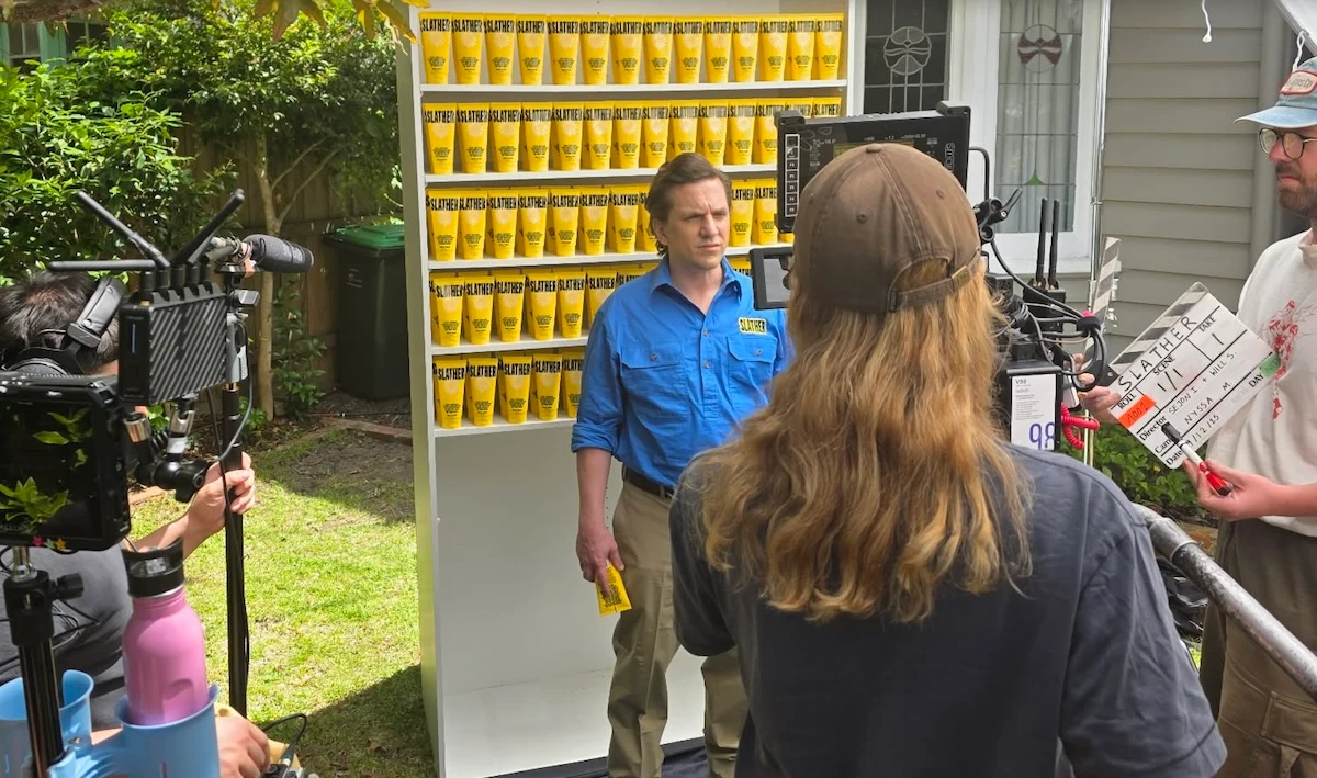 Camera crew filming a performer in a blue shirt in front of shelves of yellow SLATHER sunscreen