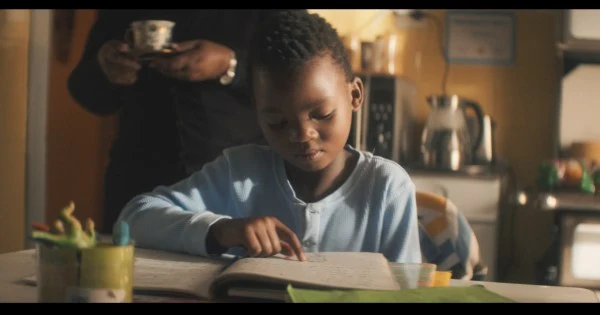 A young reader looking down at a book at a kitchen table in Cadbury’s Mrs. Mebena film